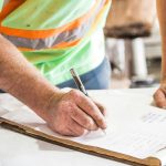 A construction worker writes notes on a clipboard at a construction site, emphasizing planning and precision.